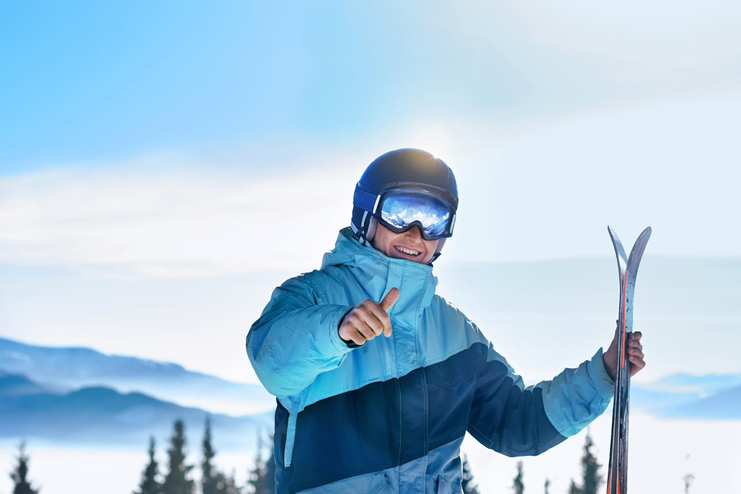 Een man met een skibril staat in de sneeuw. Hij heeft in zijn ene hand twee ski's vast en met zijn andere hand steekt hij zijn skiduim op.